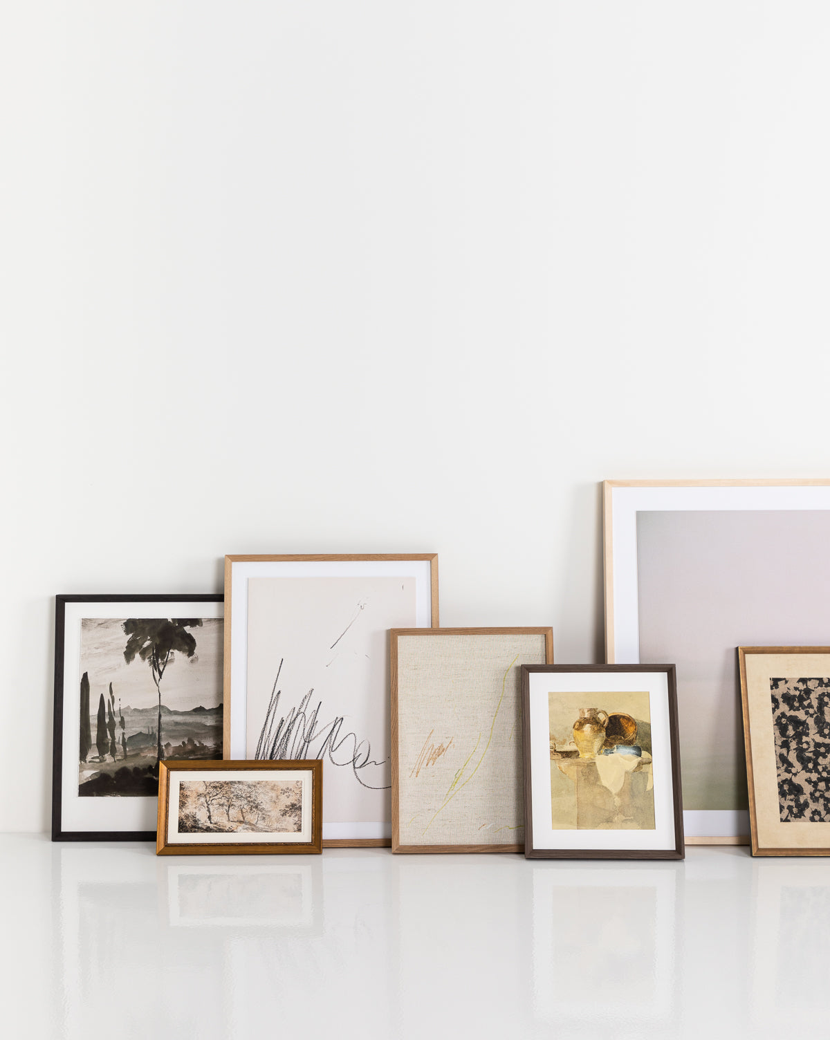 Six framed art prints, including giclee and vintage wall art, lean against a white wall on a glossy surface. The display includes landscapes, abstracts, textured images in muted tones, and the Tipped Vases print.