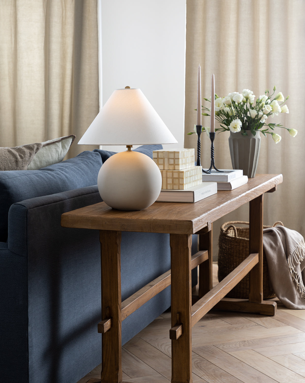 A wooden console table behind a blue sofa displays the Tess Ceramic Table Lamp, stacked books, two tall candles, and a vase of white flowers. Neutral curtains and parquet flooring complete this cozy living room setting by McGee & Co.