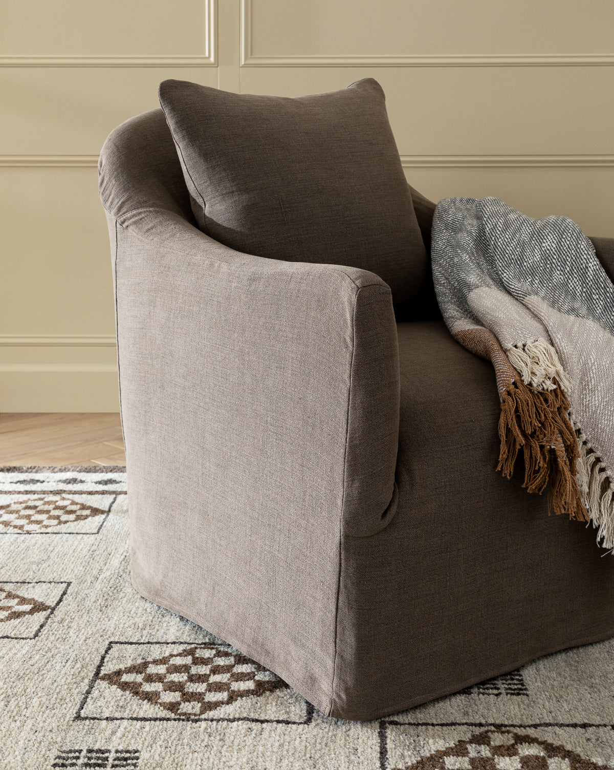 The Trudeaux Slipcover Swivel Chair (Ready to Ship), in brown upholstery with a matching cushion and Feather Blend Cushions, is draped with a gray and brown fringed throw, sitting on a patterned rug against beige paneled walls, by McGee & Co.