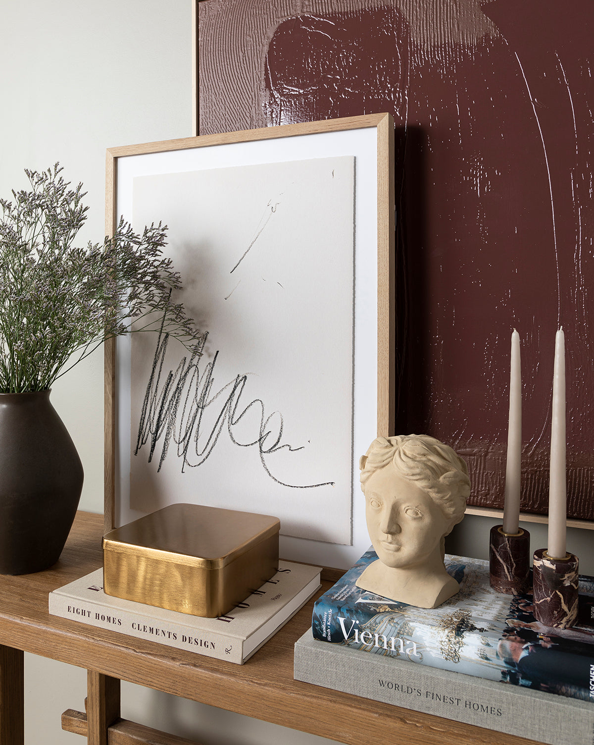 A wooden table is styled with a potted plant, abstract framed artwork, a gold box, stacked books, the Hestia Bust in composite stone, and two tall candles. Textured burgundy art hangs behind as a backdrop.