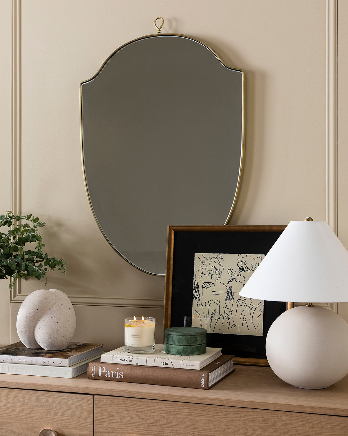 The Courtyard dresser features stacked books, a small potted plant, stone decor, a lit candle, a green box, a framed vintage illustration, a round white lamp, and an ornate wall mirror above.