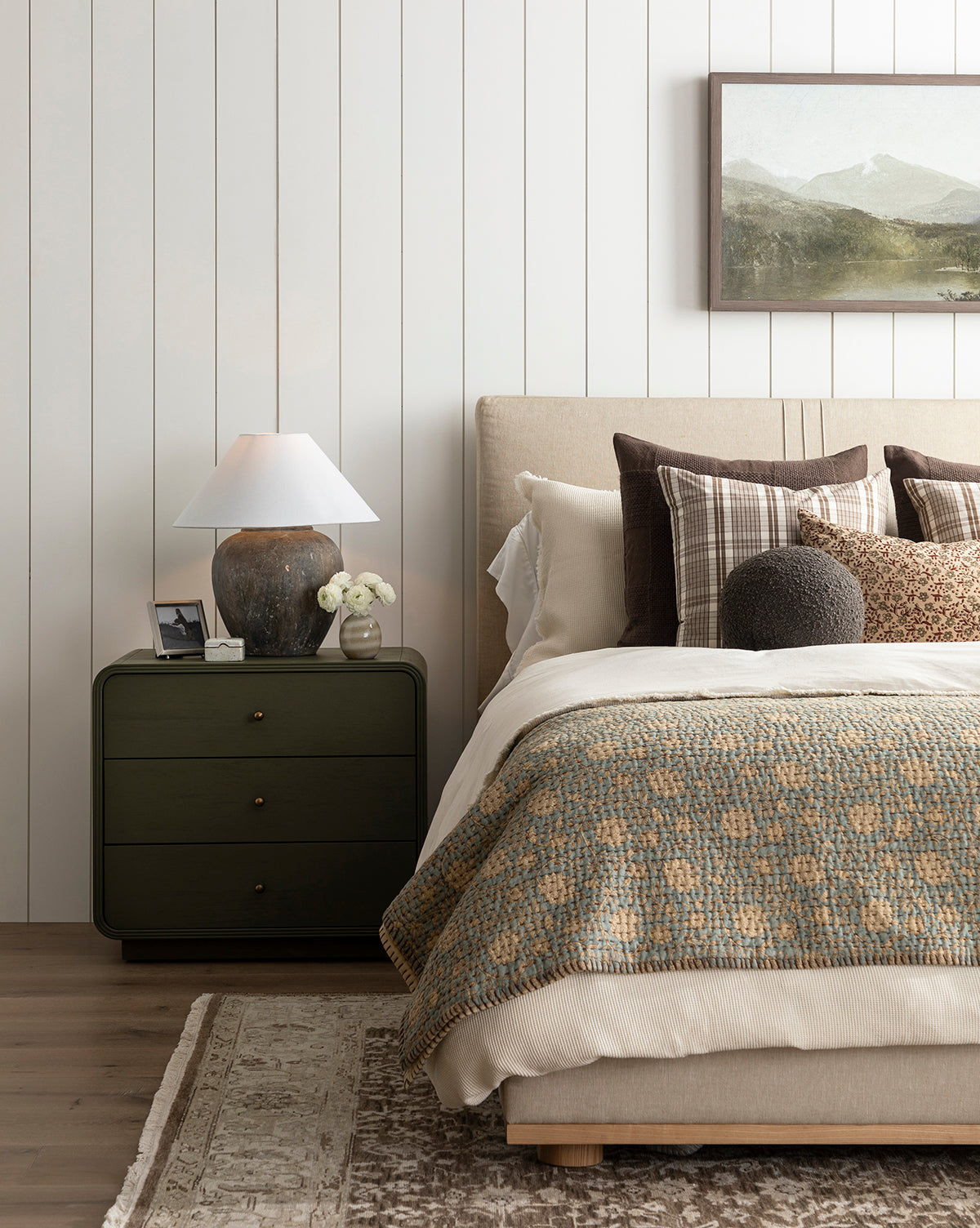 A cozy bedroom with a beige upholstered bed, plaid and textured pillows, a green knit blanket, the Gabriella Block Print Quilt, a green nightstand with a lamp and flowers, plus a landscape painting on a white shiplap wall.