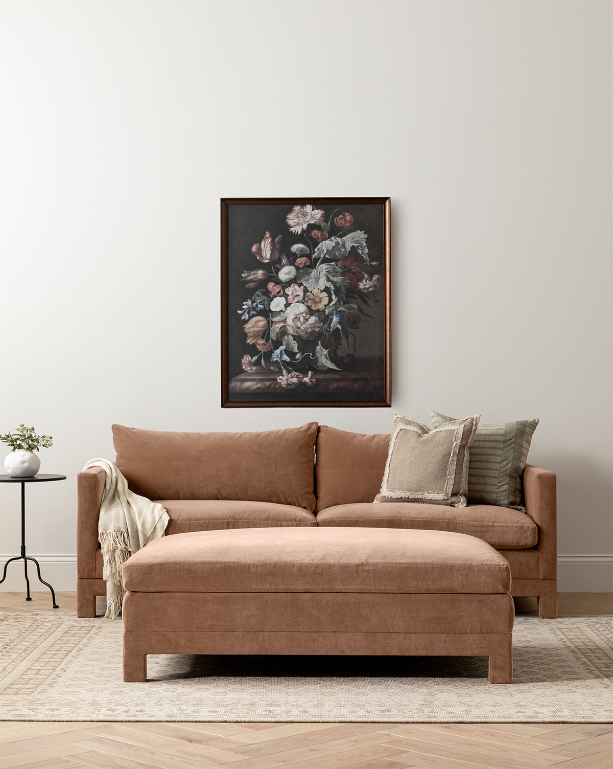 A tan Makerspalm Appoline Sofa 85 with assorted pillows and a throw anchors a minimalist living room. Above it, a floral painting hangs on the neutral wall, while a side table, plant, and beige rug complete the cozy space by McGee & Co.