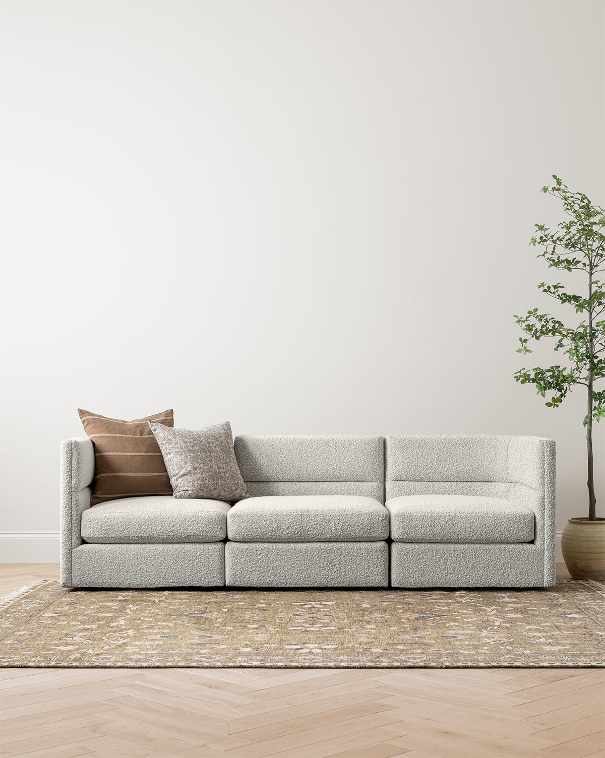 The Claremont Modular Sofa 99 by Makerspalm, featuring customizable fabric and two decorative pillows, is arranged on a patterned rug in a minimalist room with light wood flooring, cream walls, and a tall potted plant on the right, by McGee & Co.