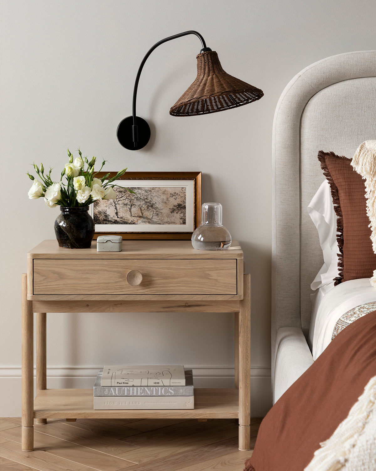 A wooden bedside table with a drawer holds a vase of white flowers, the Dutch Landscape Study giclee print in an antique gold frame, a glass of water, and a book. Above, a brown woven wall lamp is mounted next to part of a beige bed with pillows.