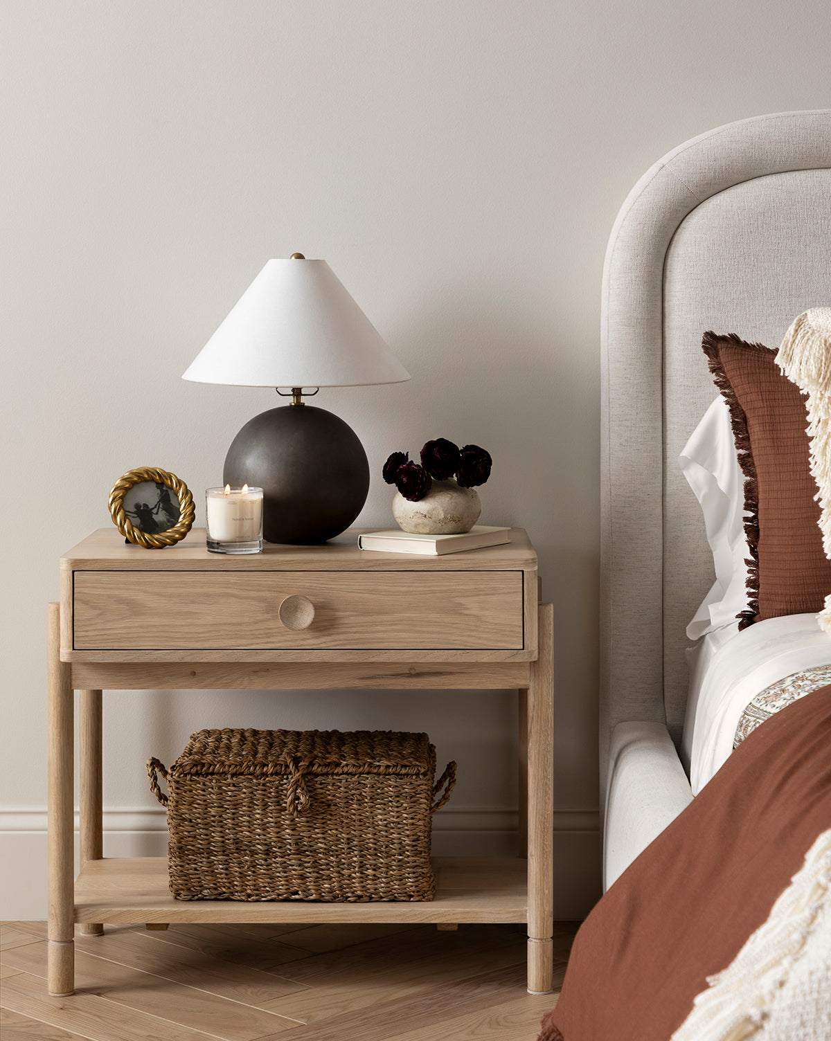 A wooden nightstand beside a bed holds a black lamp, candle, photo frame, and vase with dark flowers. The Kya Seagrass Box sits on the lower shelf. The bedding is brown and cream, by McGee & Co.