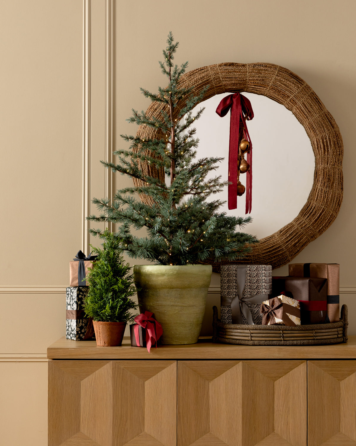 A 17" Cypress Faux Potted Tree and a tiny plant in a terracotta pot sit on a wooden cabinet with wrapped gifts. A round wicker mirror, adorned with a red ribbon and gold ornaments, hangs on the wall behind them, by McGee & Co.