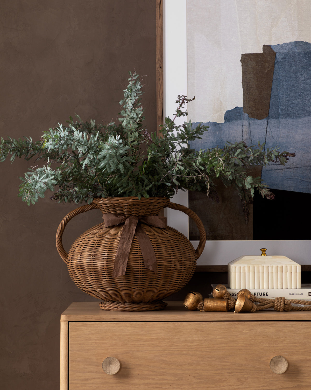 A brown wicker vase with greenery sits on a wooden dresser next to a white Plateau Lid Box, gold bells, and rope, with a framed abstract artwork hanging on the wall behind.