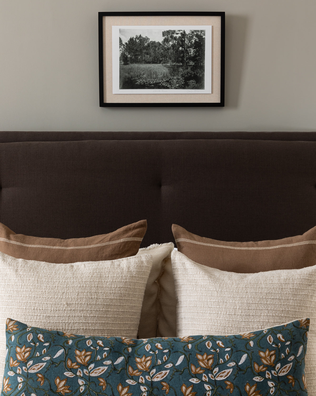 A neatly made bed with beige and brown pillows, plus a blue floral cushion, sits against a taupe wall. Above the bed, Muted Florida Pines by Carol Haymon, a black-and-white giclee print, is framed and centered.