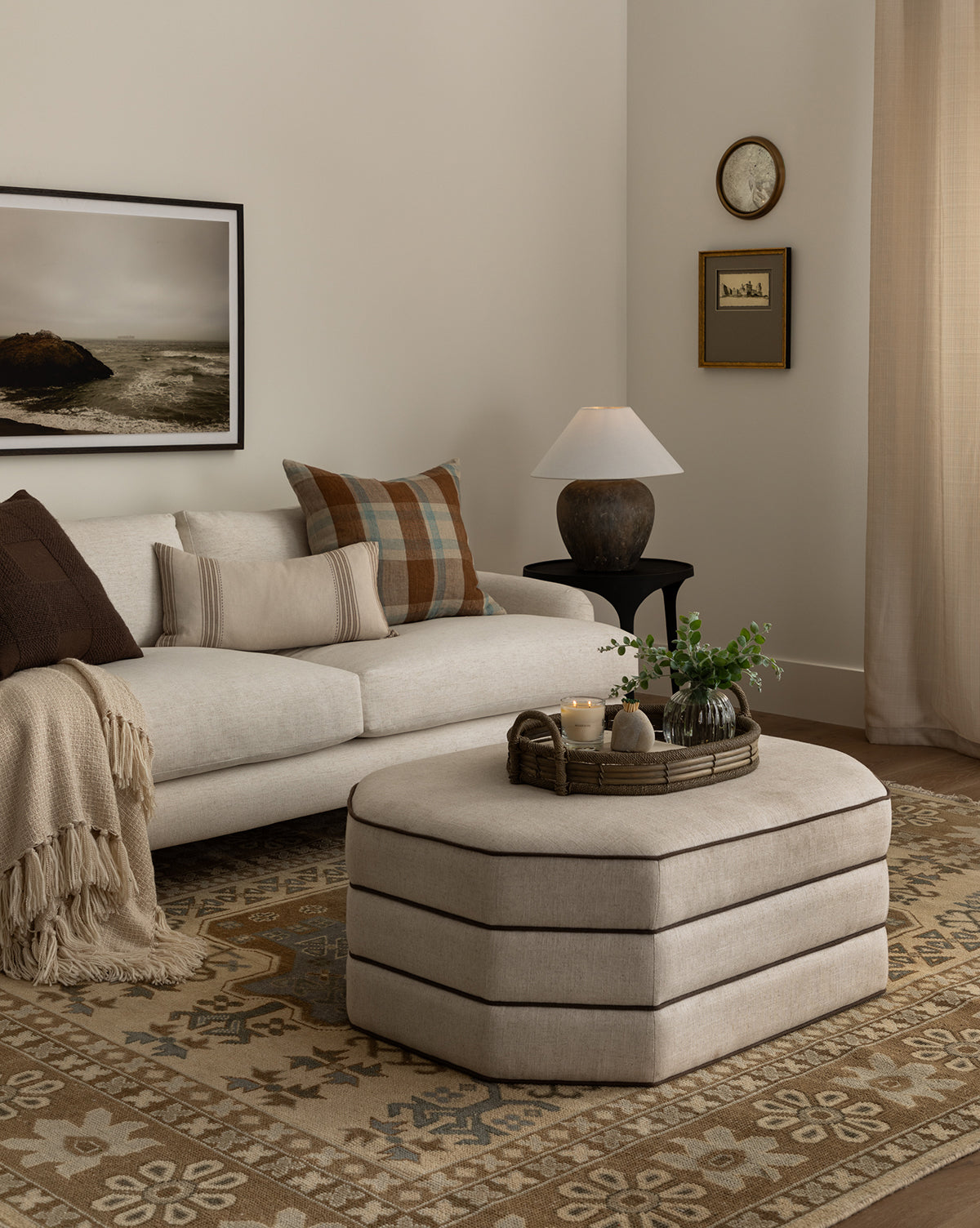 A cozy living room featuring a light beige performance linen sofa with neutral pillows, a plaid cushion, and a tan throw. The Makerspalm Rafferty Ottoman displays candles and a plant, while artwork hangs above a patterned rug—McGee & Co.