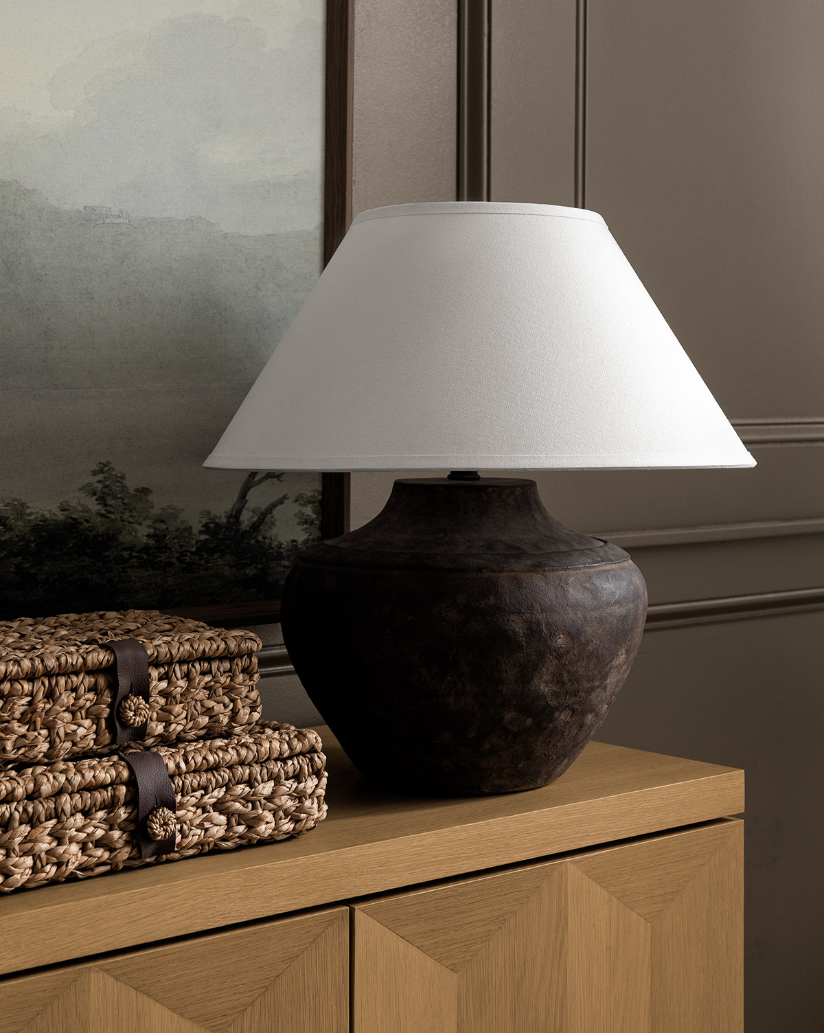 The Gannon Table Lamp, crafted from dark ceramic with a fabric shade, sits on a wooden sideboard beside stacked woven baskets, framed by a landscape painting and paneled wall in the background, by McGee & Co.
