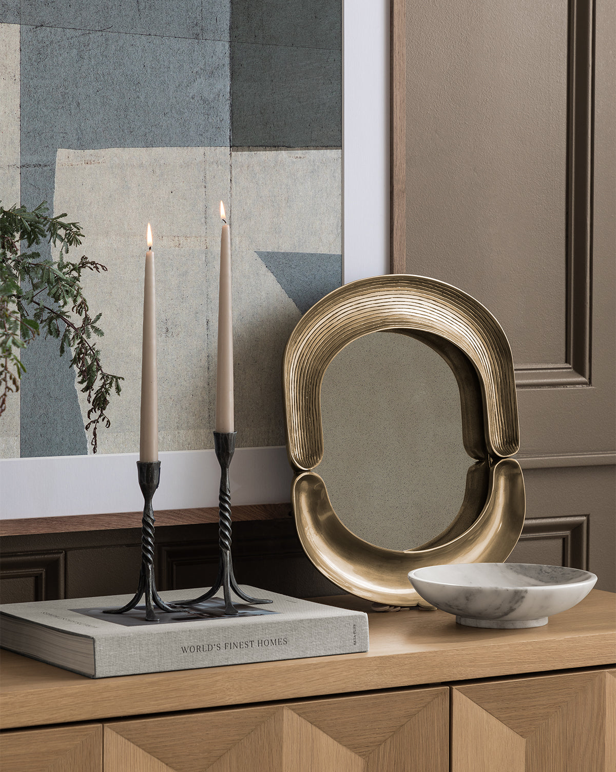 A wooden surface displays two lit candles in black holders on a book, a gold tray, and the Arabesco Marble Bowl, set before a taupe wall and modern artwork.