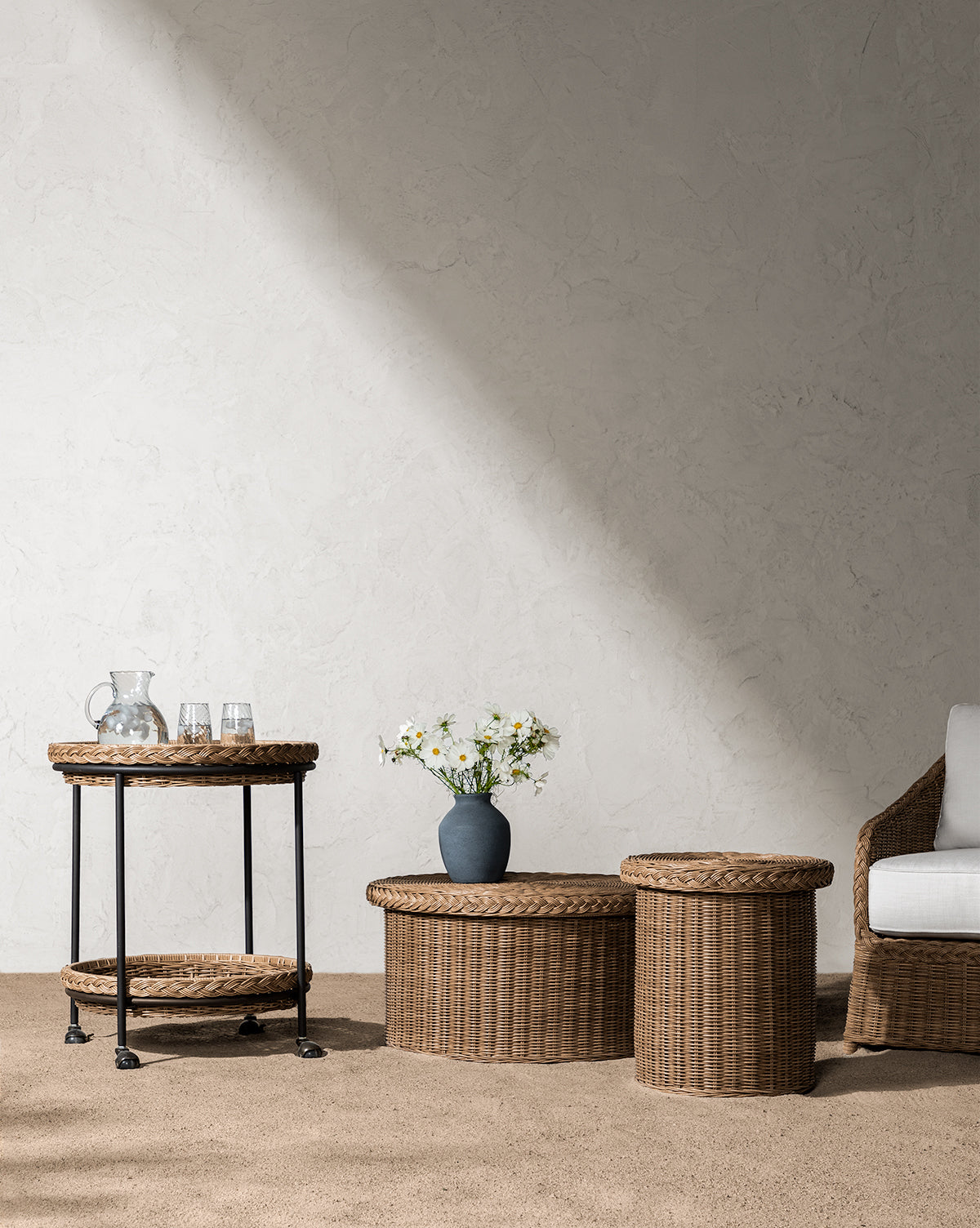 A minimalist room showcases the weather-resistant Haviland Side Table, a bar cart with a glass pitcher and glasses, two wicker tables, a vase of white flowers, and a white cushioned chair against a textured wall lit by sunlight, by McGee & Co.
