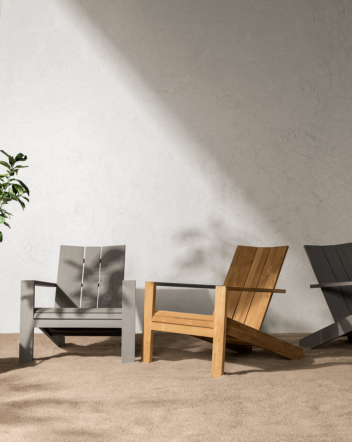 A Triby Gray Teak Outdoor Lounge Chair sits on a beige carpet against a textured light wall, with sunlight casting shadows and a green plant partially visible to the left, by McGee & Co.