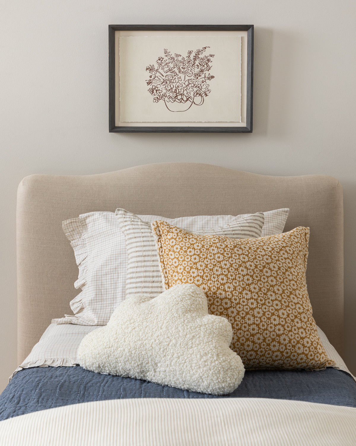 A neatly made bed with striped, ruffled, mustard floral, and white cloud-shaped pillows sits below the Tea Cup of Flowers giclee print in a wood frame on antique white watercolor paper, set against a light-colored wall.