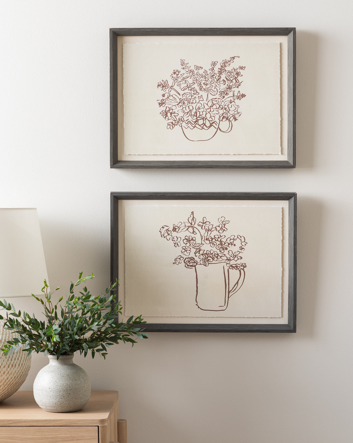 The artwork Pitcher of Flowers, a framed botanical still life line drawing in a charcoal wood grain frame, hangs above a wooden table with a potted plant and lamp on a light-colored wall.