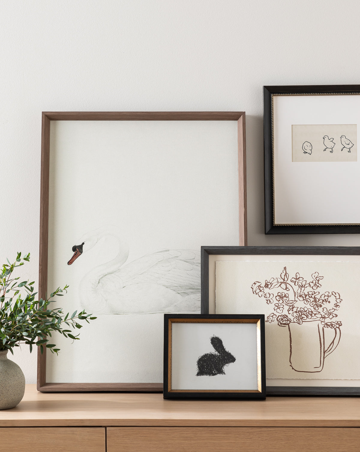 A wooden sideboard displays framed artwork: a swan drawing, chicks sketch, the Pitcher of Flowers botanical still life, and a black rabbit silhouette. A leafy plant in a vase sits on the left.