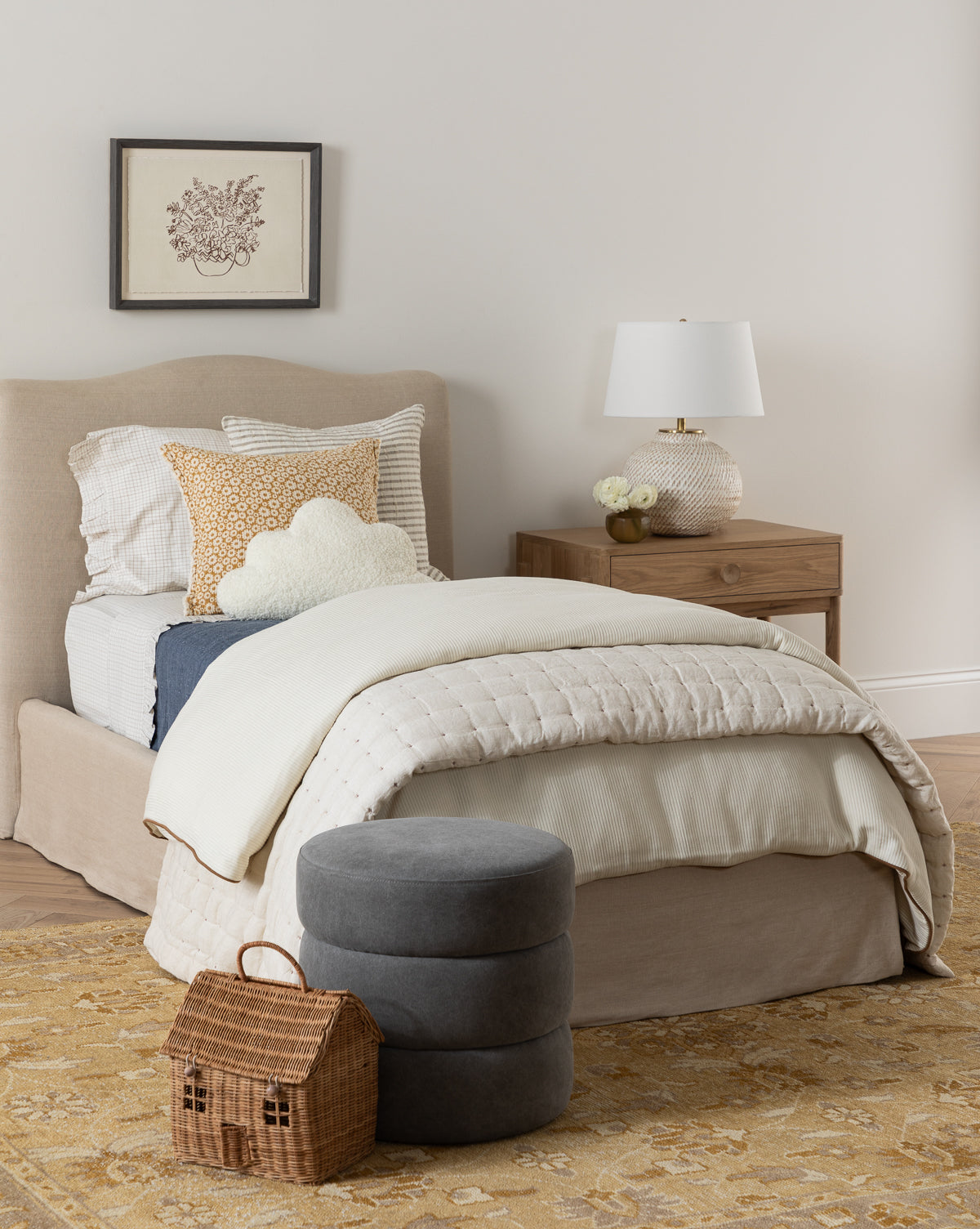 A cozy bedroom featuring a beige bed, white and patterned pillows, the blue Talcott Quilt, a wooden nightstand with a white lamp, a framed floral picture, gray ottomans, a wicker house basket, and a beige patterned rug.