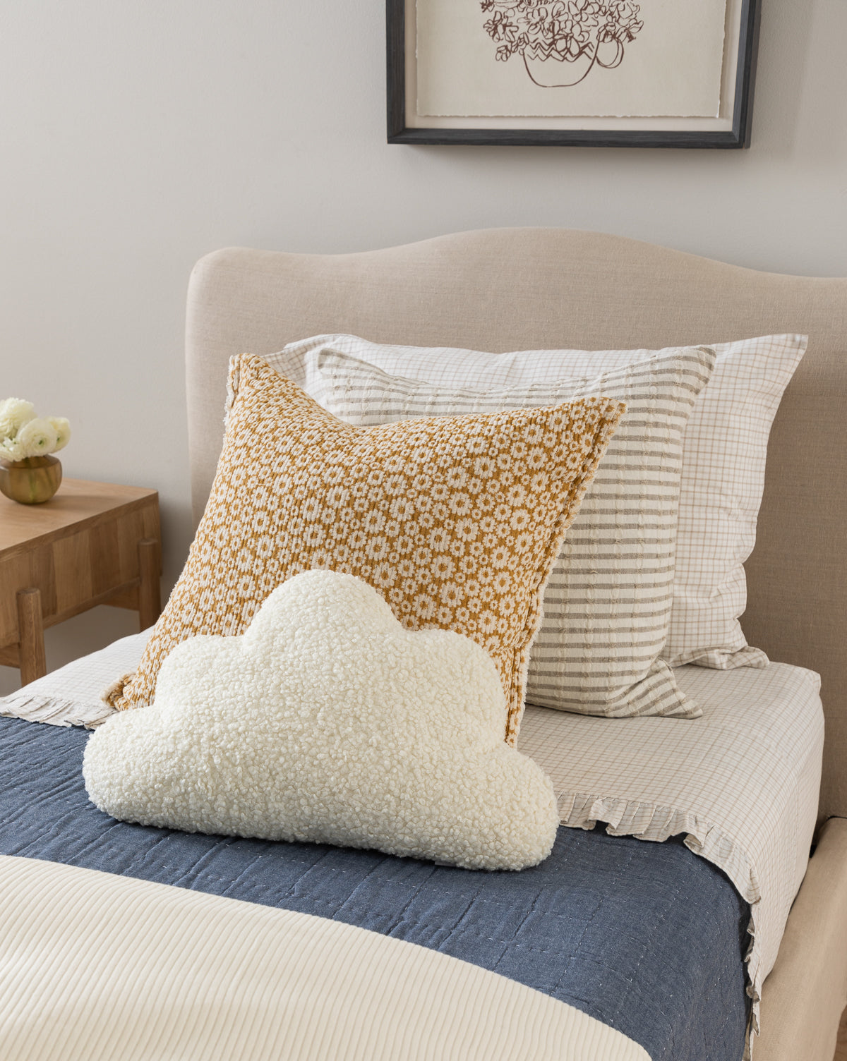 A neatly made bed features the Fitzgerald Kids Quilt in stripes, accented by a mustard floral pillow and a white cloud-shaped pillow. A wooden nightstand with flowers sits beside the bed, with framed artwork above the headboard.