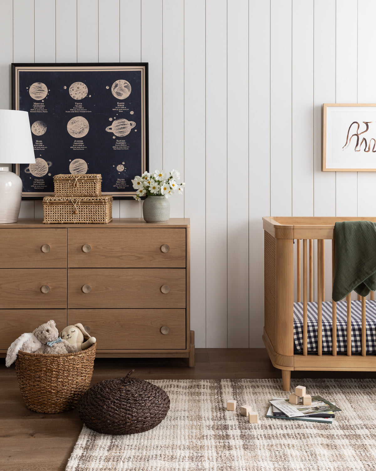A cozy nursery with a wooden crib, dresser, plaid rug, woven basket of stuffed animals, and the Nine Planets Diagram wall art. The room has a neutral palette and vintage-inspired decor for a charming childrens space.