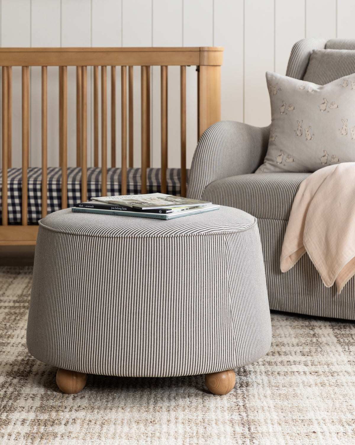 A cozy living room featuring the Barnaby Ottoman with stripes, a gray armchair and pink blanket, a wooden crib, and a patterned rug. Books sit on the ottoman amid decor in neutral and soft tones, by McGee & Co.