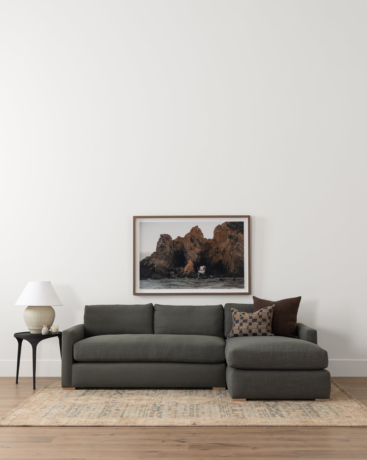 A contemporary living room featuring the Community Macy Upholstered Chaise Sectional 101 in dark gray, patterned throw pillows, a side table with a white lamp and vase, a light rug, and a framed coastal rock photo on a white wall, styled with McGee & Co.