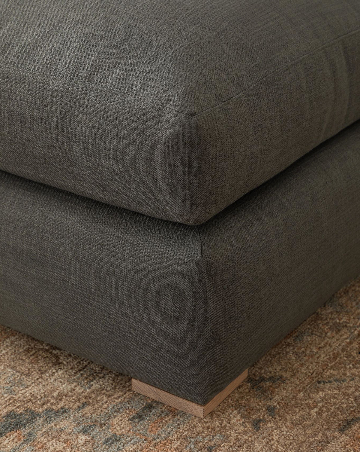 Close-up of the Macy Upholstered Chaise Sectional 101 by Community in dark gray, featuring a plush cushion and low square wooden legs, set on a beige and brown textured rug for a modern look. McGee & Co.
