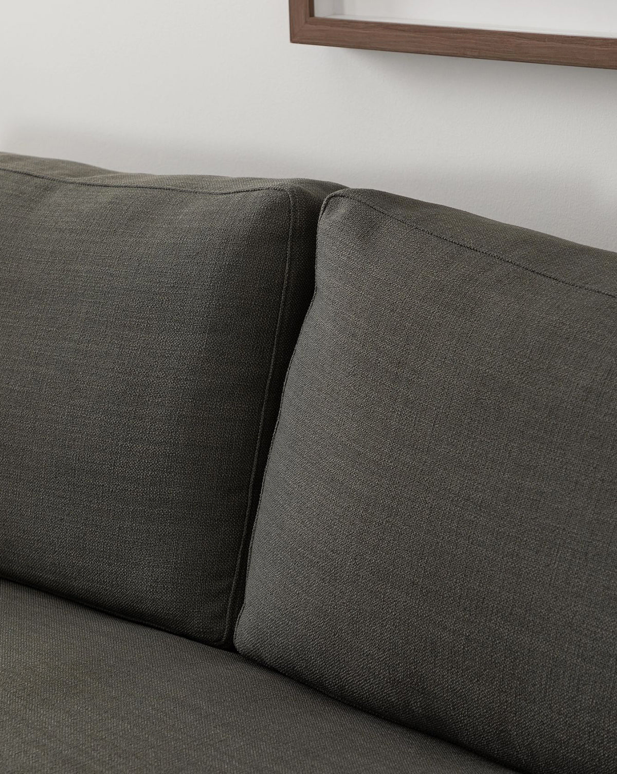 Close-up of the Macy Upholstered Chaise Sectional 101 by Community in dark gray, featuring two seat cushions and a partial picture frame on a white wall in the background. McGee & Co.