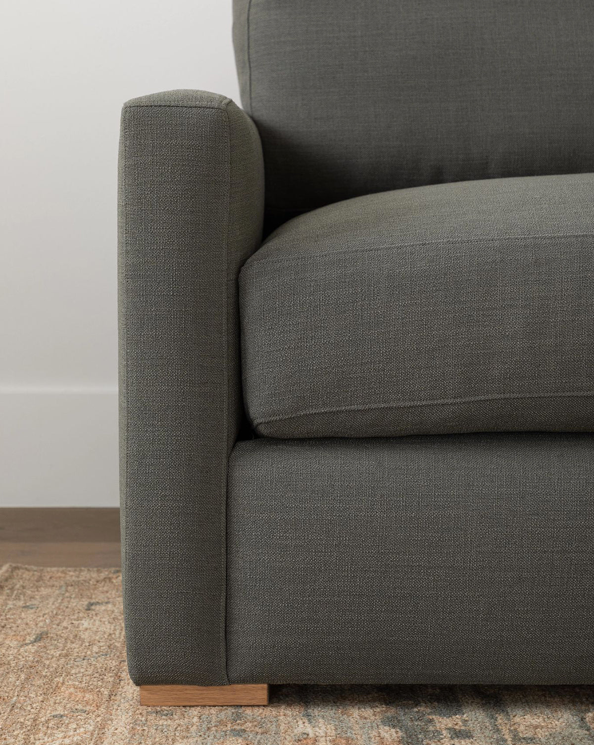 Close-up of the Macy Upholstered Chaise Sectional 101 by Community in dark gray, highlighting its armrest, seat cushion, and wooden leg, with a glimpse of a patterned rug and wooden flooring underneath. McGee & Co.