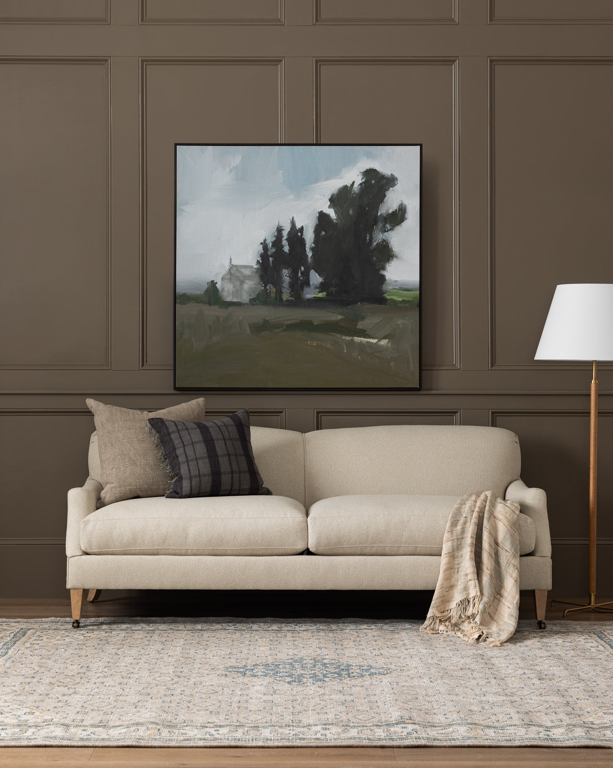 A modern living room showcases the Rowley Sofa 85 by Rowe Fine Furniture, Inc. with customizable fabric, plaid and solid pillows, a plaid throw, floor lamp, gray-blue rug, and landscape painting on a taupe paneled wall—finished with touches from McGee & Co.