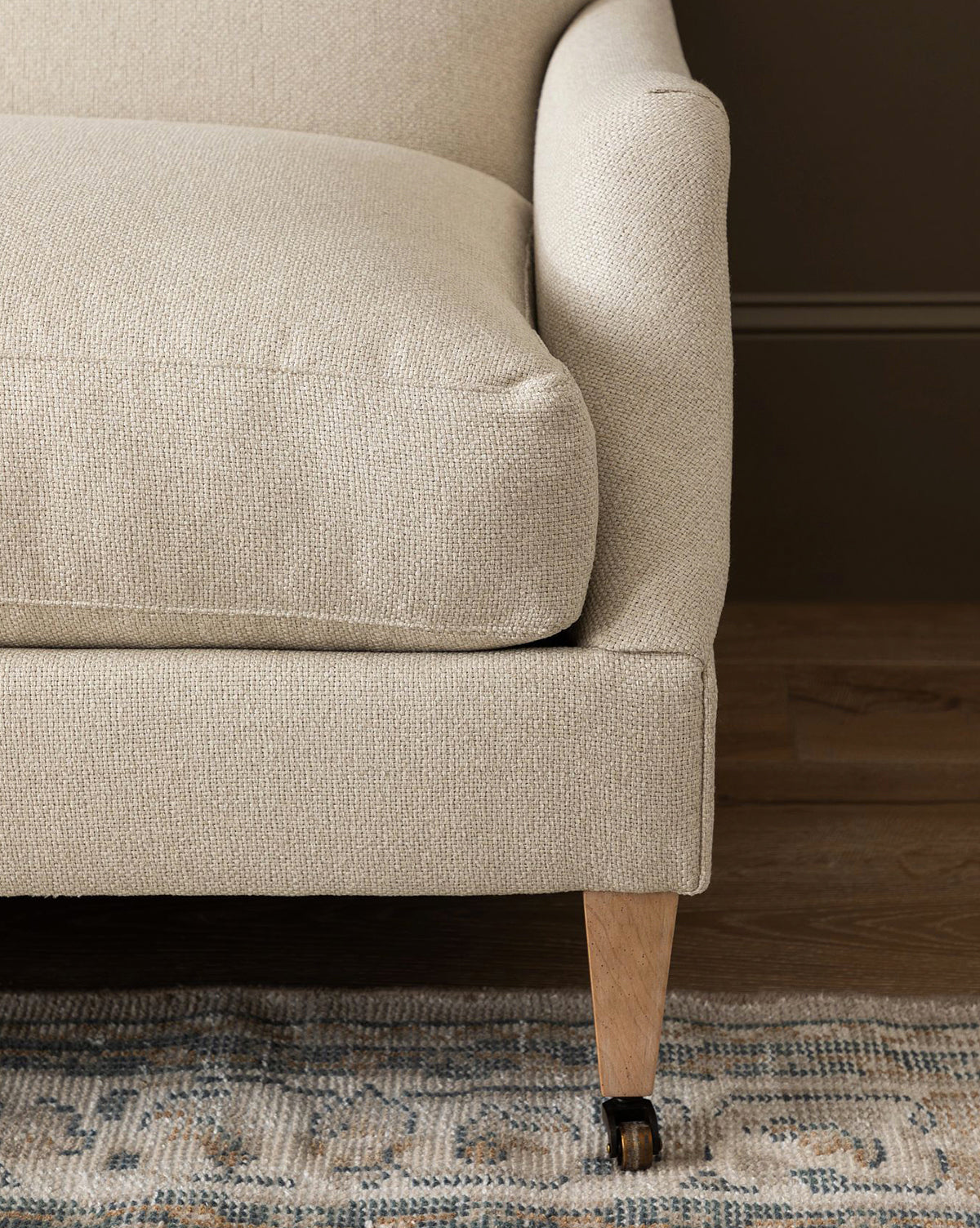 A close-up of the Rowley Sofa 85 by Rowe Fine Furniture, Inc., featuring a hand-crafted plush seat cushion, wooden leg with a caster wheel, and patterned rug on wood flooring. Choose from customizable fabric options to fit your style. McGee & Co.