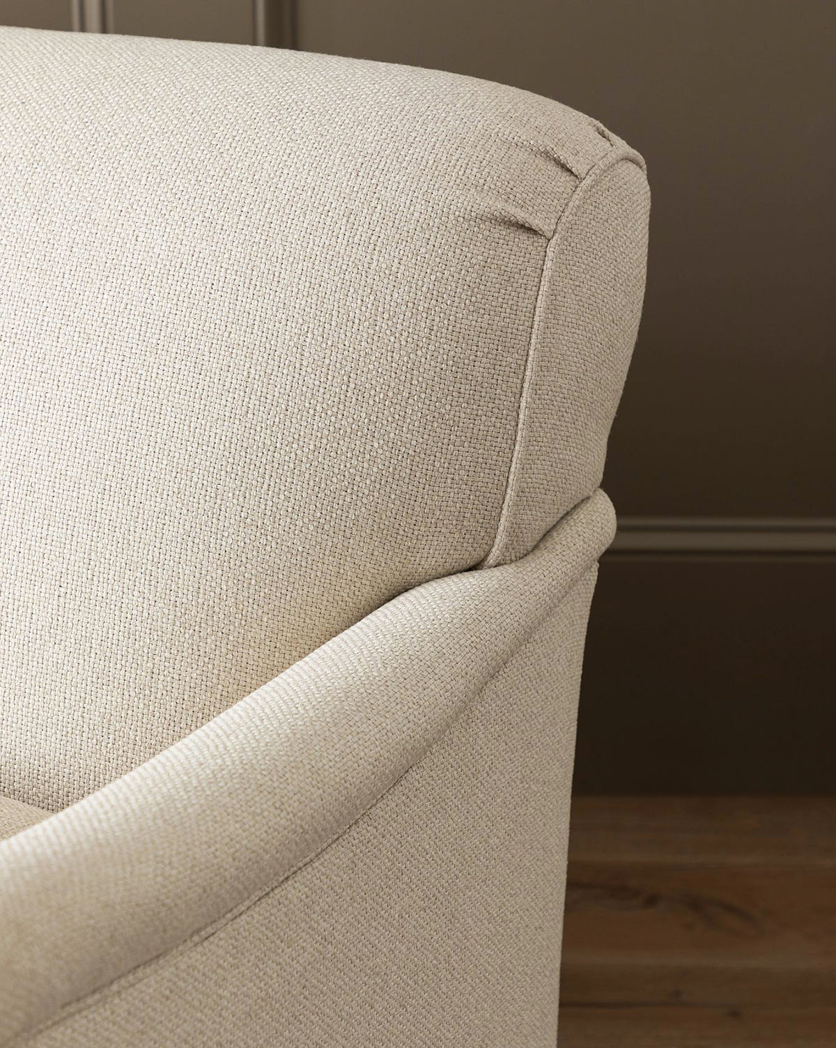 Close-up of the upper corner of the light beige Rowley Sofa 85 by Rowe Fine Furniture, Inc., featuring textured fabric and a curved armrest against a brown wall and wooden floor. Explore customizable fabric options for this hand-crafted piece. McGee & Co.