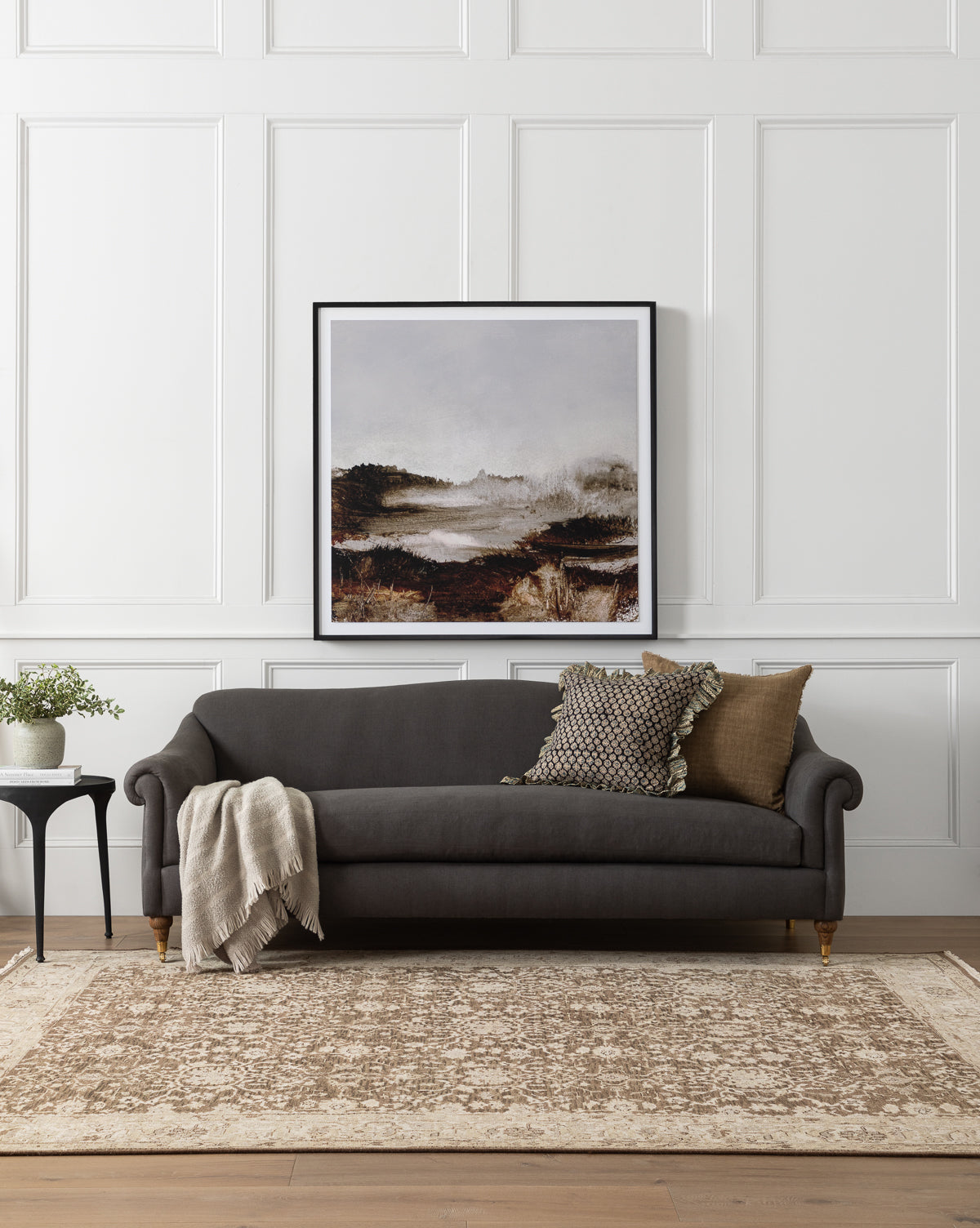 A modern living room exudes luxurious comfort with the Community Hildegard Sofa 90 in dark gray, assorted pillows, a soft blanket, side table with a potted plant, beige patterned rug, and a large framed landscape painting on white paneled walls by McGee & Co.