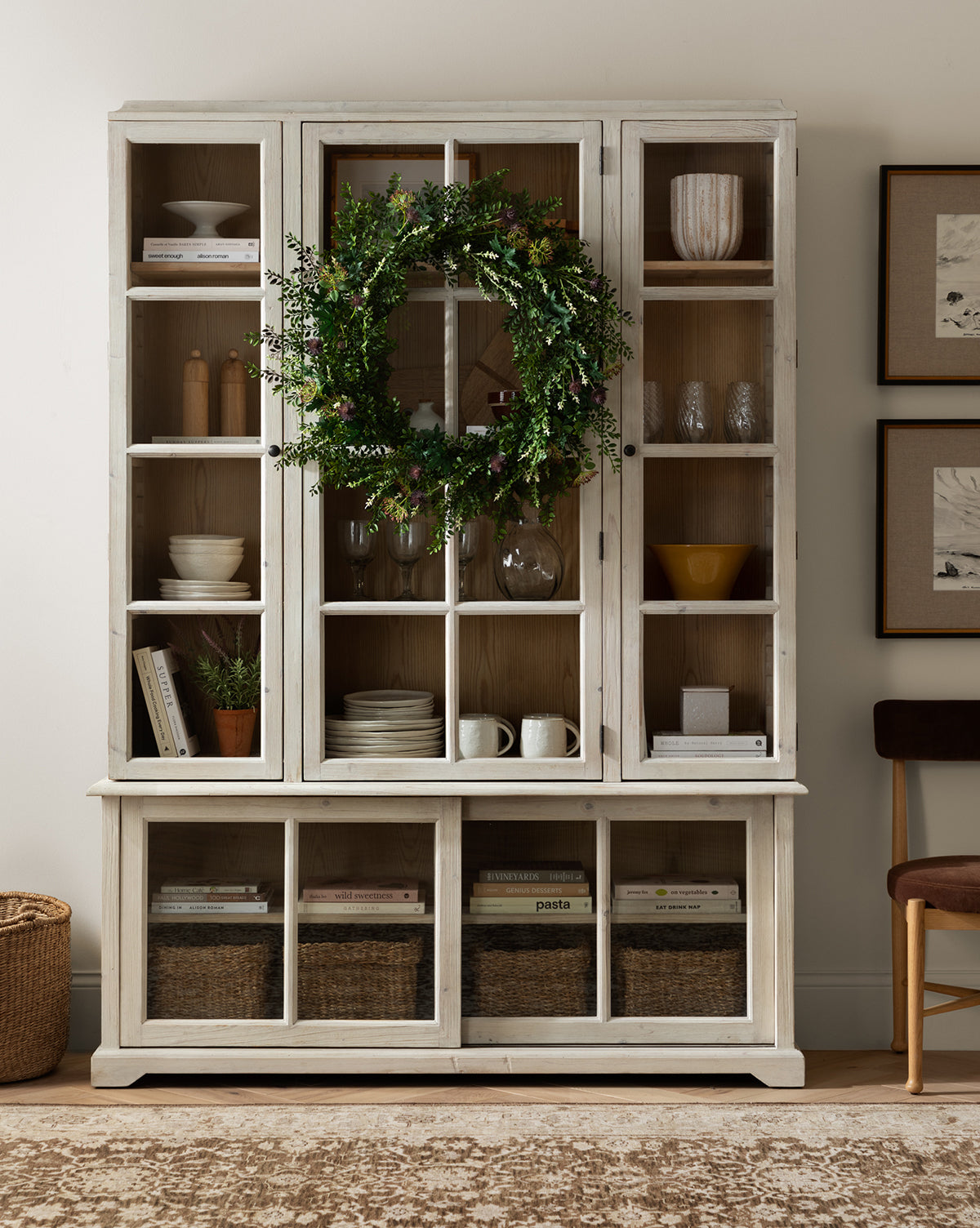 A white wooden cabinet with glass doors displays dishes, books, and glassware. A Faux Summerset Wreath hangs on the center door. Woven baskets line the lower shelves, while framed art and a brown chair are placed nearby.