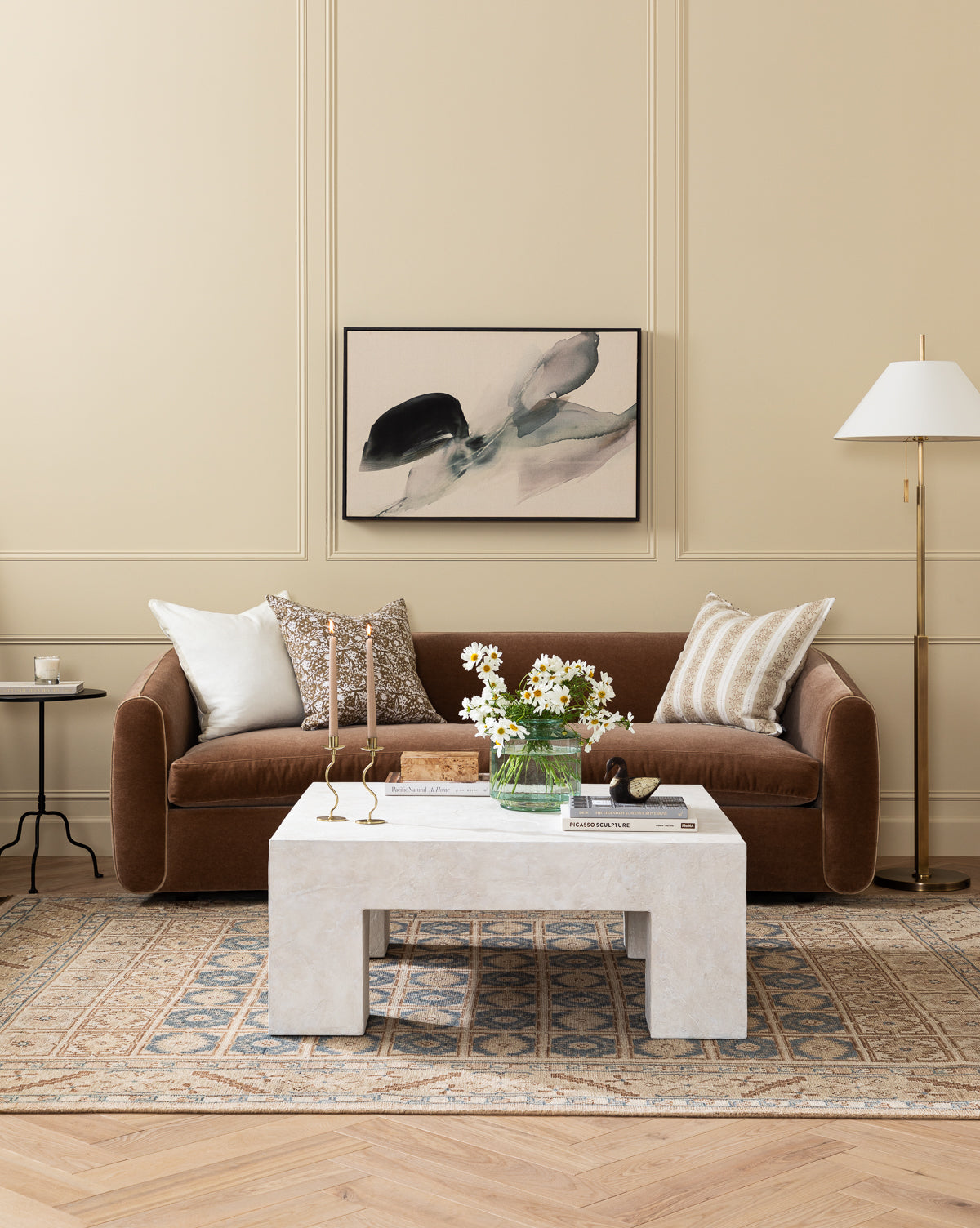 A modern living room with a brown sofa, patterned and white pillows, a marble coffee table topped with flowers and candles, the Clic Floor Lamp with an off-white linen shade and pull chain, abstract art on the wall, and a patterned rug over wood floors by McGee & Co.