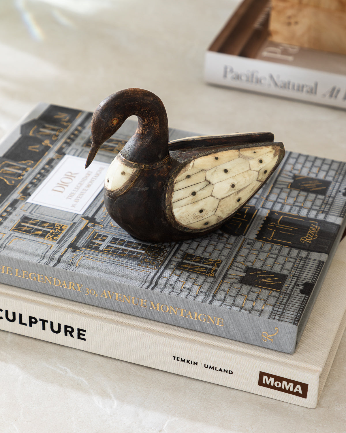 The Zella Swan Object, a vintage-inspired dark wooden sculpture with bone inlay, rests atop art and design books like DIOR, THE LEGENDARY 30, and MoMAs SCULPTURE.