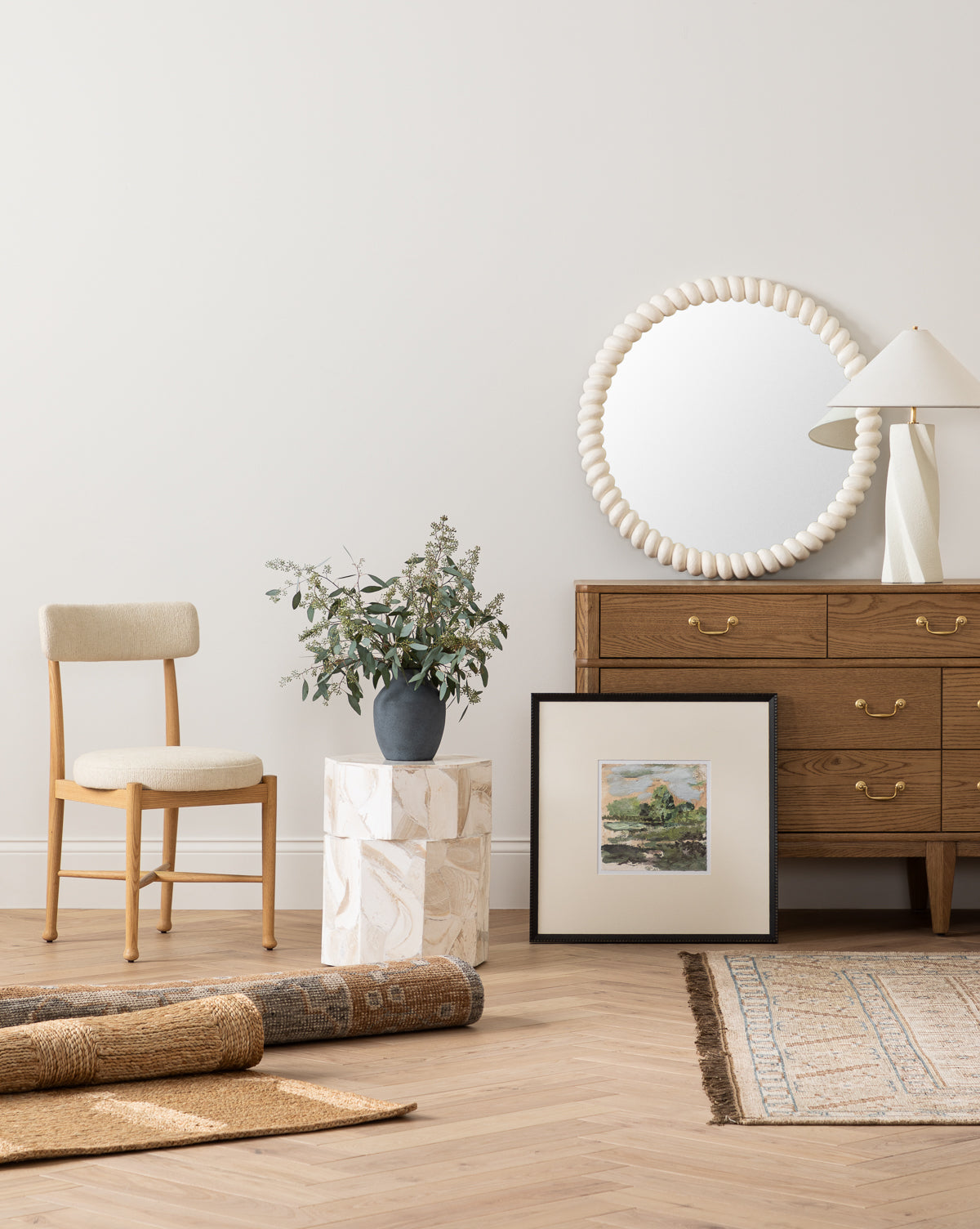 A minimal modern room featuring a wooden chair, dresser, Norina Mirror, table lamp, framed art, potted plant atop a marble pedestal, and rolled rugs on herringbone wood flooring—ideal for fans of subtle coastal style, by McGee & Co.
