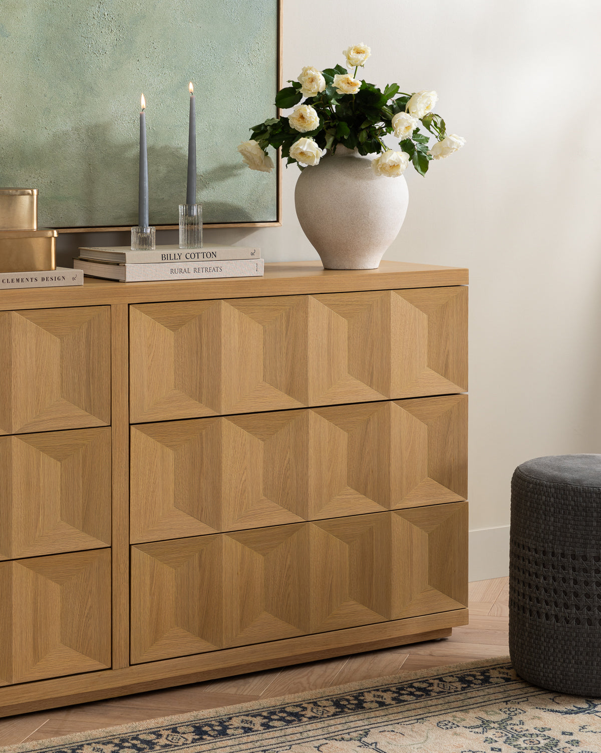 A wooden dresser with a geometric pattern stands against the wall. On top are two lit candles, stacked books, and a Krissan Vase filled with white roses. Part of a painting and a patterned rug can also be seen, by McGee & Co.