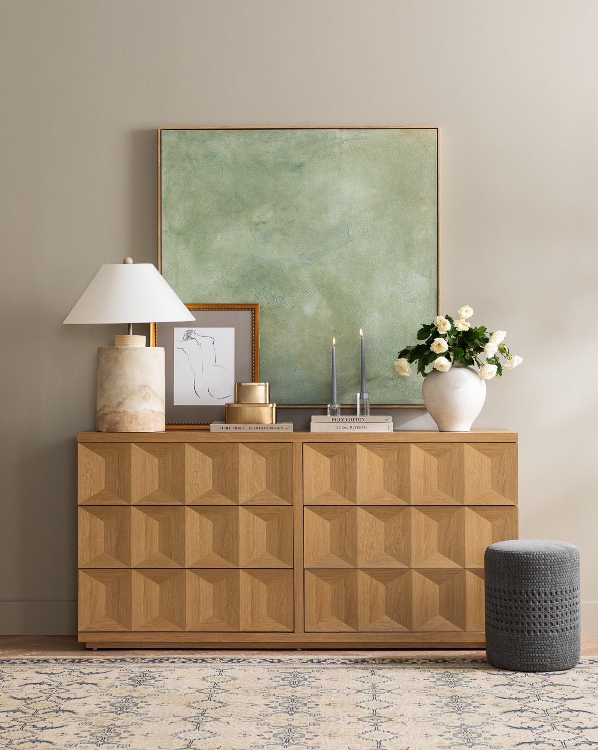 A geometric wooden sideboard is topped with a Samala Lamp, framed art, candles, a white vase of flowers, and vintage accents. Above hangs a large green abstract painting on a neutral wall, with a gray pouf placed close by, by McGee & Co.