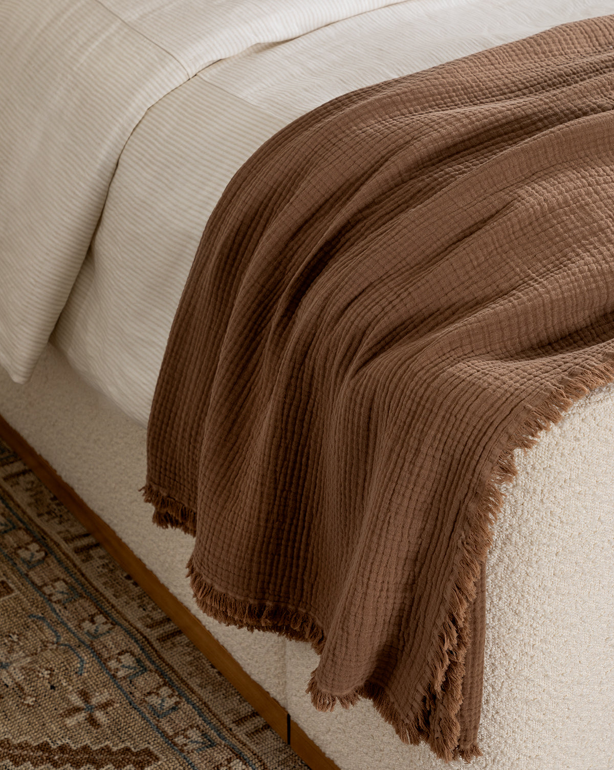 The Marinella Oversized Waffle Throw, featuring a textured brown cotton design with fringed edges, drapes over the side of a cream-colored bed beside a patterned rug on the floor.