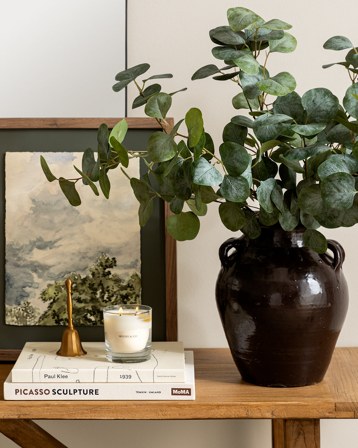 A dark ceramic vase with leafy green branches sits on a wooden table next to a lit candle, brass bell, art book, and the Head in the Clouds art print in a walnut frame leaning against the wall.