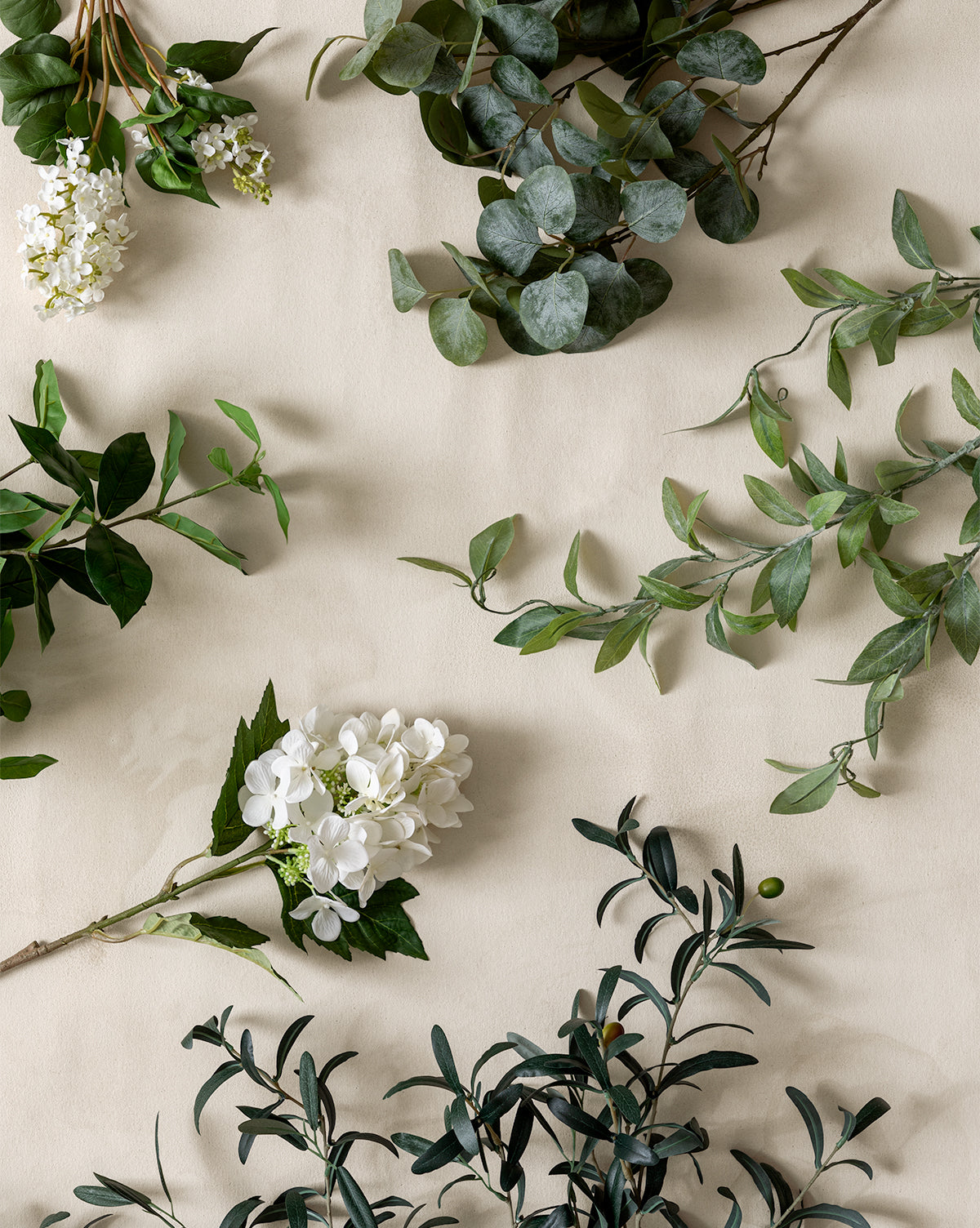 A Faux White Lilac Stem, along with green eucalyptus and olive branches, is arranged on a light beige fabric background for a scattered, natural look.