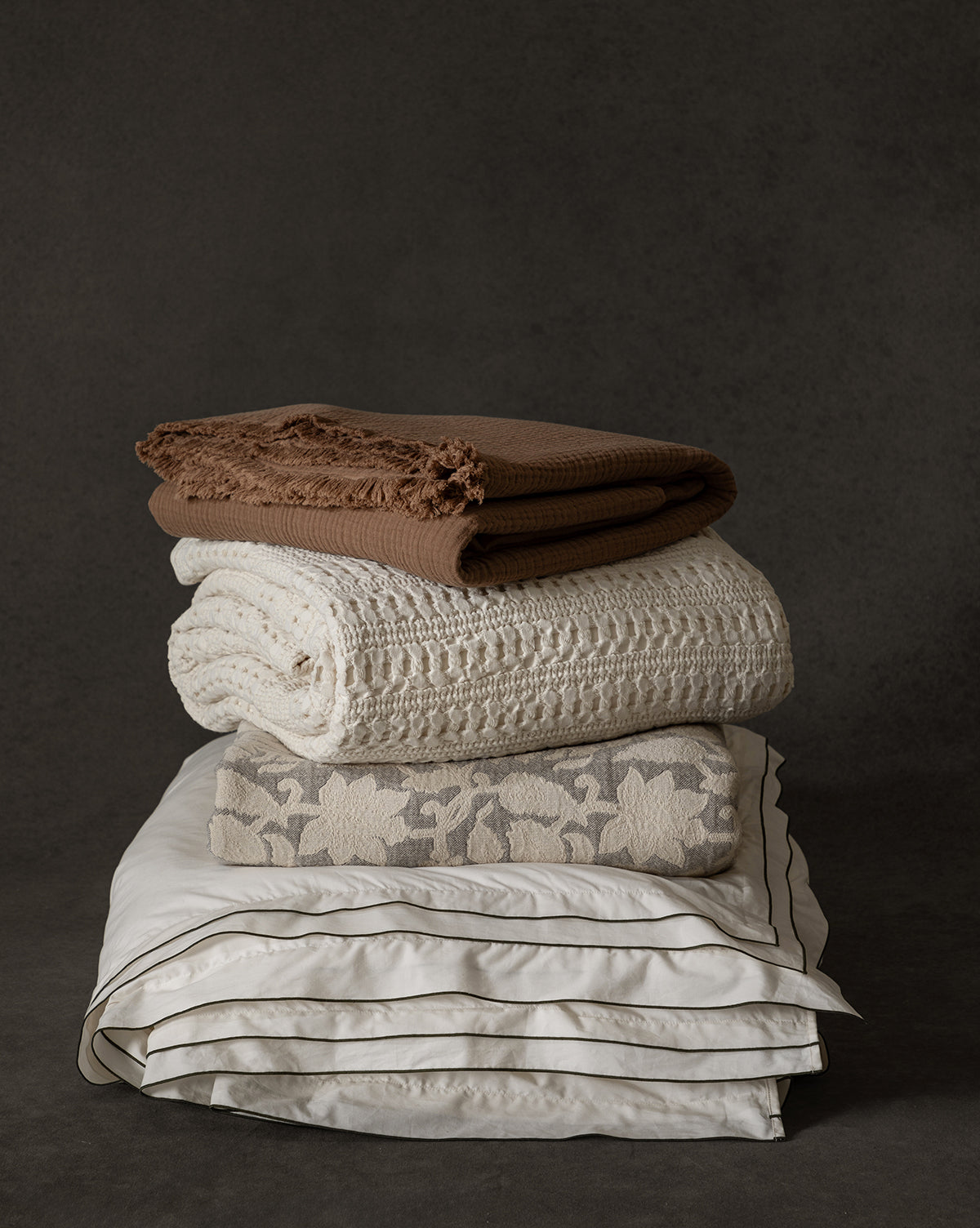 A neatly stacked pile of neutral-toned blankets features the Marinella Oversized Waffle Throw, showcasing cozy waffle weave texture and soft cotton alongside other throws with various textures and patterns, set against a dark background.
