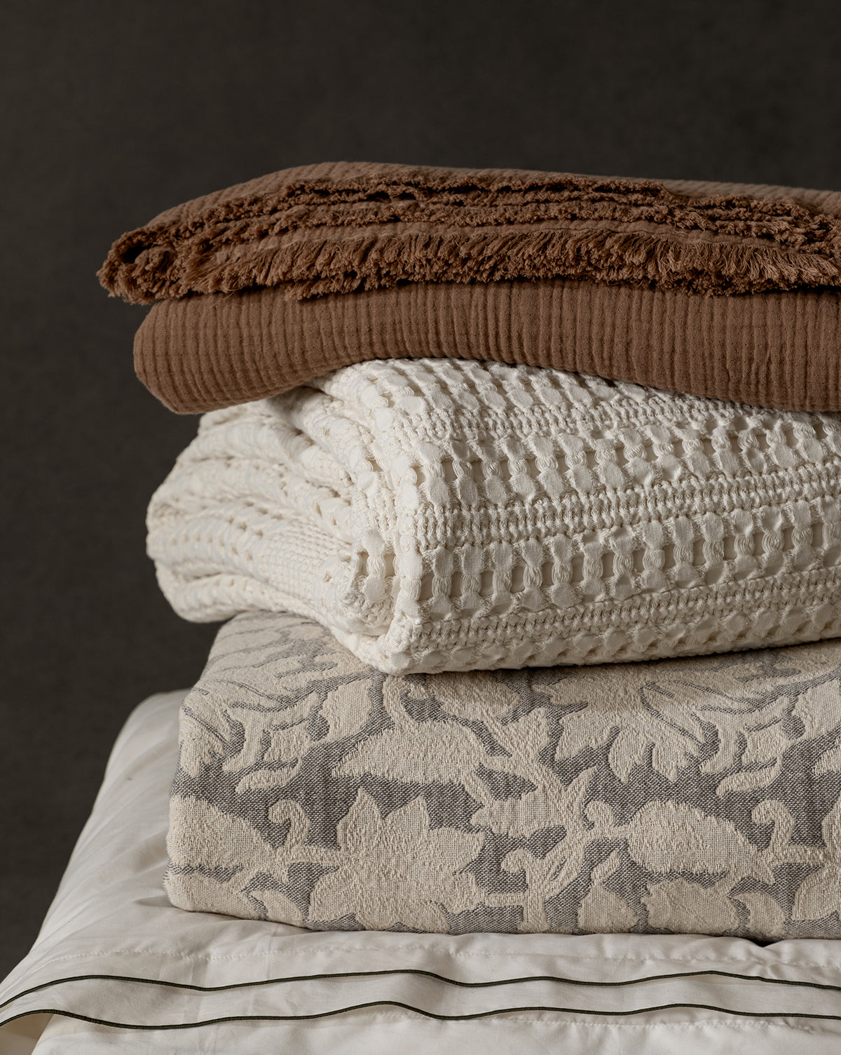 A neatly stacked pile of folded blankets in various textures and neutral tones, featuring the Carnel Waffle Blanket—crafted from 100% cotton in Portugal—alongside floral and crocheted designs, set against a dark background.