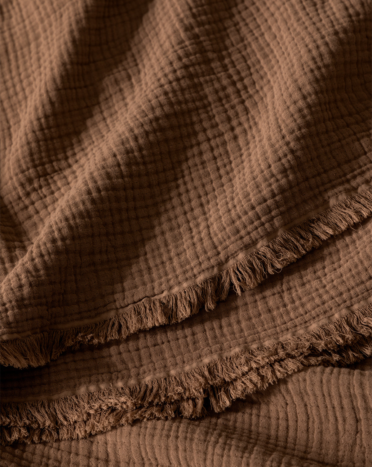 A close-up of the Marinella Oversized Waffle Throw, featuring a brown, textured cotton design with a crinkled, grid-like waffle pattern and fringed edges, draped in soft, overlapping folds.