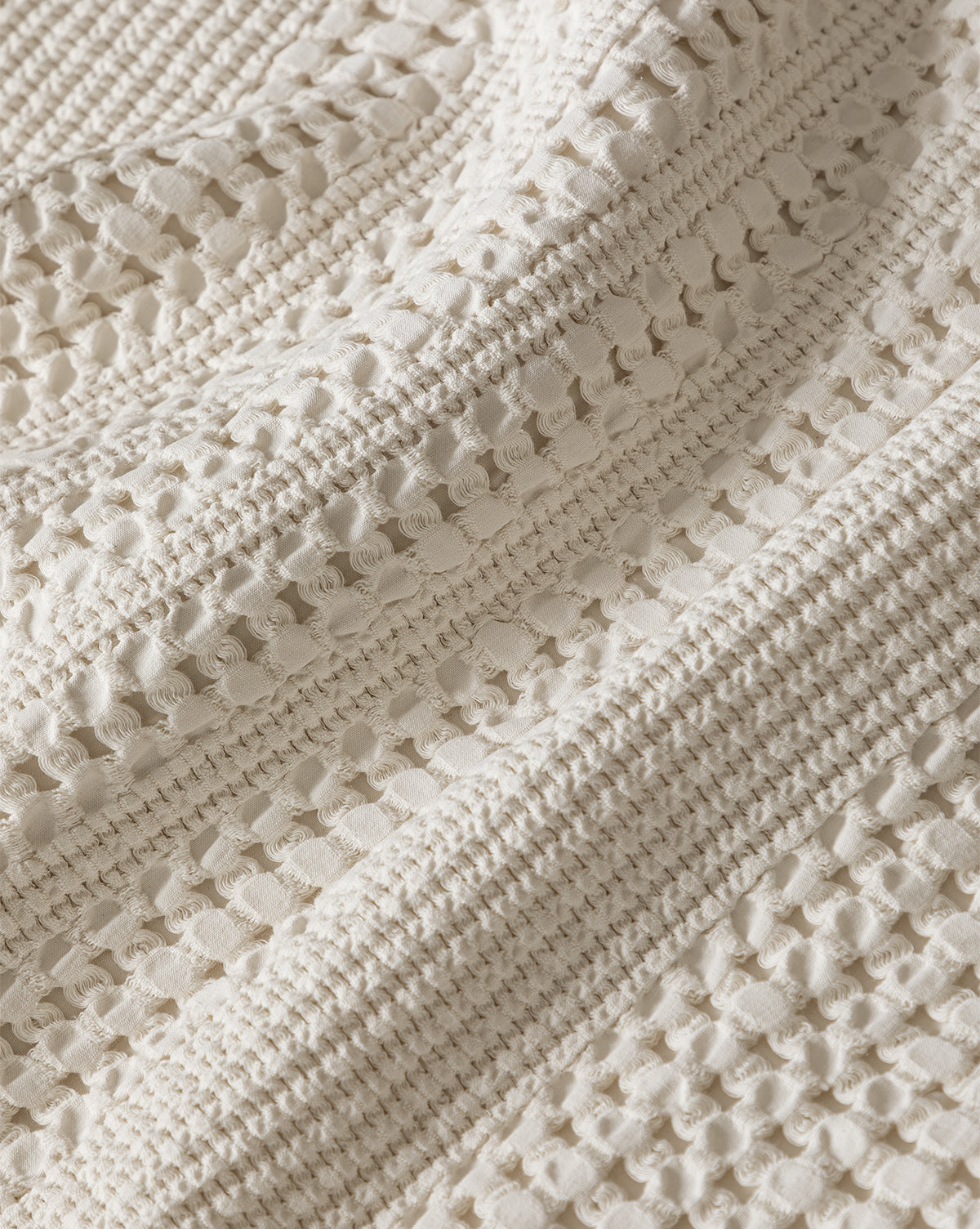 Close-up of the Carnel Waffle Blanket, crafted in Portugal from 100% cotton. It features a cream color, soft textured surface, and a repeating waffle-knit pattern with delicate holes and raised stitches.
