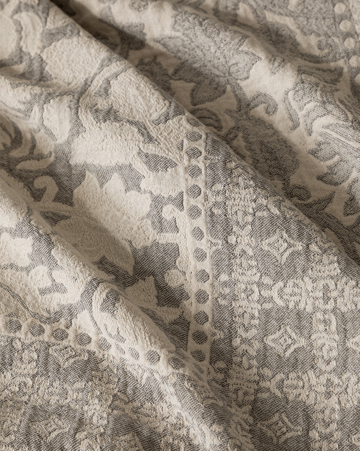 Close-up of the Floral Matelasse Coverlet, featuring a textured cotton surface with ornate floral and geometric patterns in soft beige and gray tones, highlighting its elegant folds and subtle shadows.