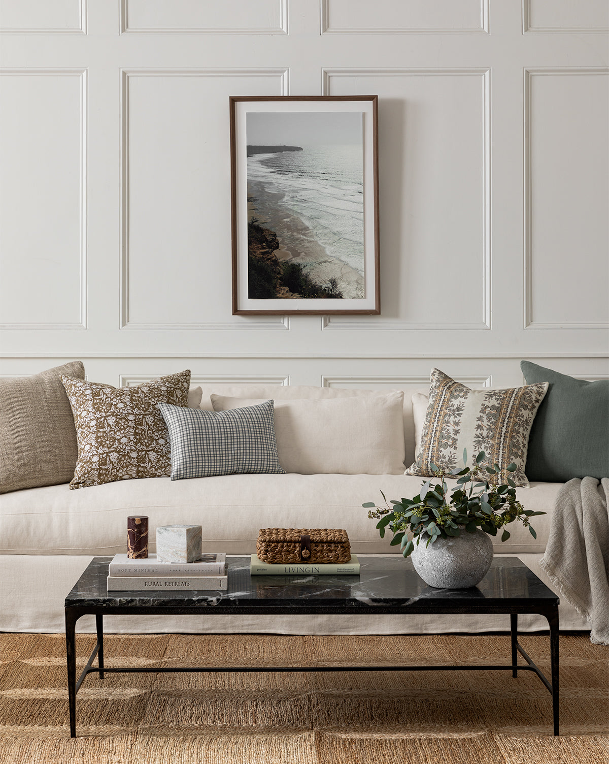 A neutral-toned living room includes the Wilhelmina Classic Slipcover Sofa (85-110) with patterned pillows, a black marble coffee table topped with books and a plant, a woven rug, and a framed coastal photo on a paneled white wall, by McGee & Co.