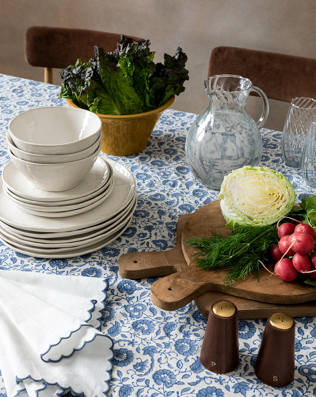 A table with a blue and white floral tablecloth features stacked white dishes, a bowl of leafy greens, a glass water pitcher, napkins, salt and pepper shakers, and the Boylan Bread Board with lettuce, radishes, and dill.