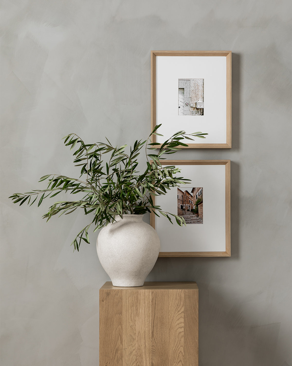 The Valldemossa white ceramic vase with green leafy branches sits on a wooden pedestal, while two giclee prints in natural woodgrain frames hang on a light gray textured wall behind it.
